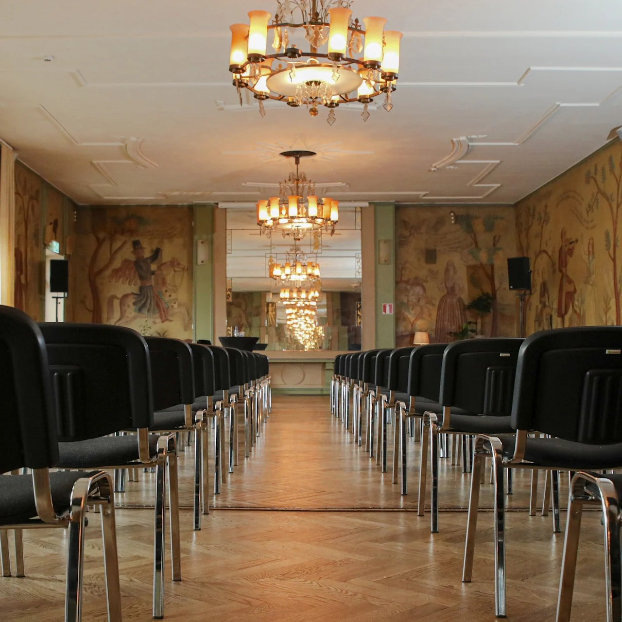 Rows of black chairs facing an ornate mirror in an elegantly decorated room with chandeliers and murals. Walls have floral and human motifs, creating a refined atmosphere.