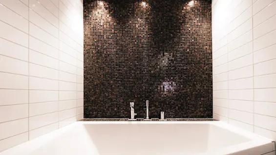 Et hvidt badekar med moderne design står mod en væg med små, mørke mosaikfliser. Kromarmaturer er synlige, omgivet af hvide flisebelagte vægge.