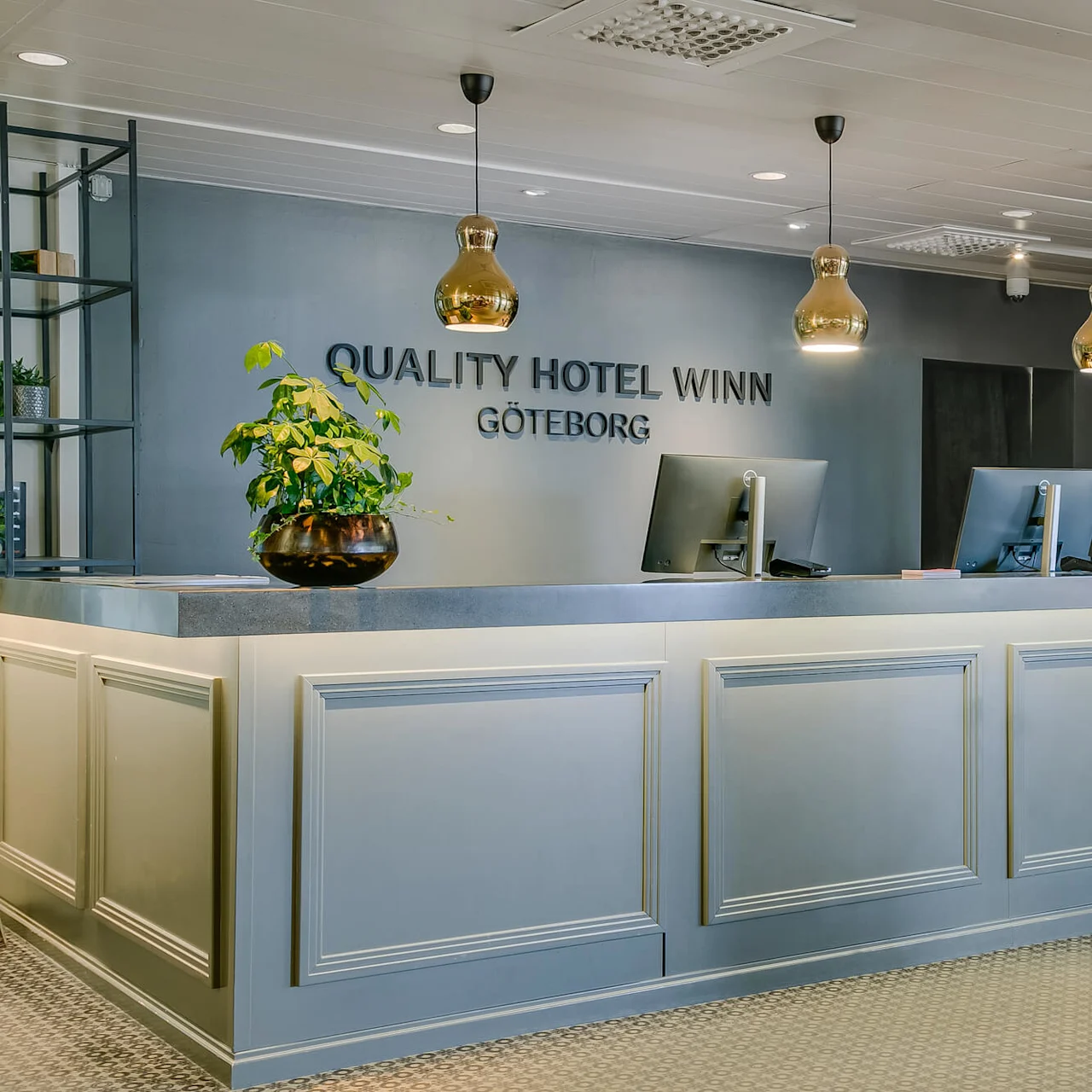 Receptionen med computere og planter under loftslamper i en hotellobby. Teksten på væggen lyder "QUALITY HOTEL WINN GÖTEBORG."