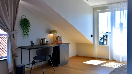 Ett ljust, mysigt rum med snedtak, skrivbord, stol och ett fönster med utsikt. Solljus strömmar in genom en dörr med skira gardiner.