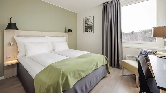A neatly made bed with white pillows and a green blanket, in a modern room with light-colored walls, a large window with a curtain, and a small desk and chair.