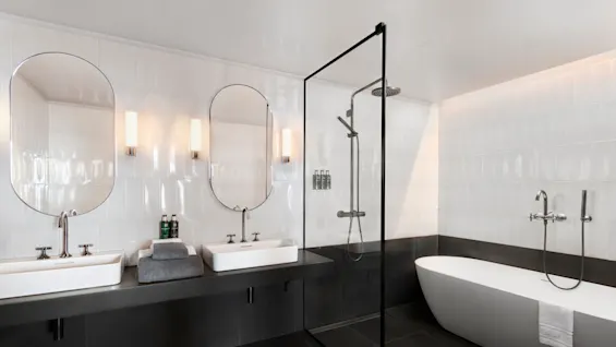Ett modernt badrum med två handfat och speglar, en dusch med glasvägg och ett fristående badkar. Rummet har elegant svart och vit kakel med minimalistiska armaturer.
