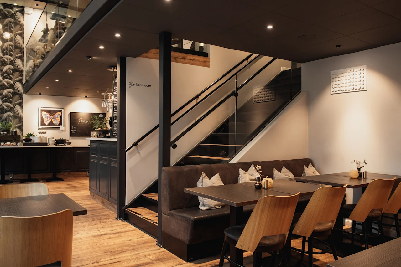 Et moderne hotelinteriør med spiseborde, båse, en morgenmadsdisk og en trappe. Et skilt indikerer Toilet og God morgen.