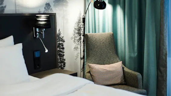 A cozy hotel room features a neatly made bed, an upholstered armchair with a pillow, and a bedside lamp. Dark curtains and a wallpaper with tree designs complete the tranquil setting.