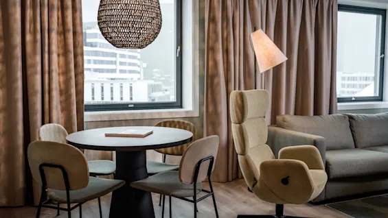 Et moderne oppholdsrom med et spisebord med stoler, en komfortabel lenestol og en sofa, med utsikt over byen utenfor vinduet.