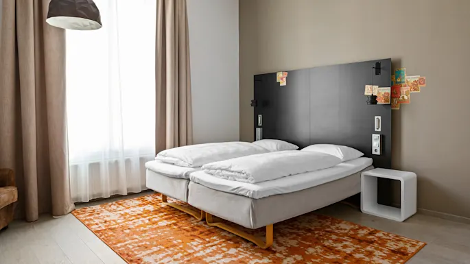 A double bed with white bedding sits on an orange patterned rug. A black headboard features vintage-style postcards. Beige curtains hang beside a window, and a geometric ceiling lamp is overhead.