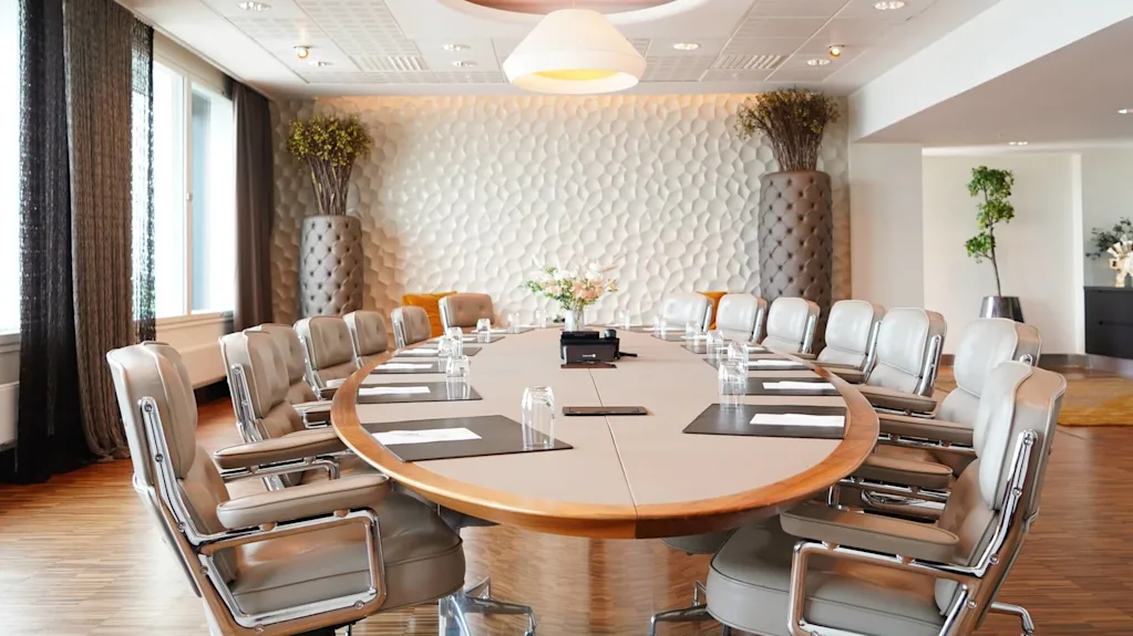 Ett stort ovalt konferensbord omgivet av läderstolar, placerat i ett modernt rum med strukturerade väggar, stora krukor och fönster med gardiner. Papper och glas finns på bordet.