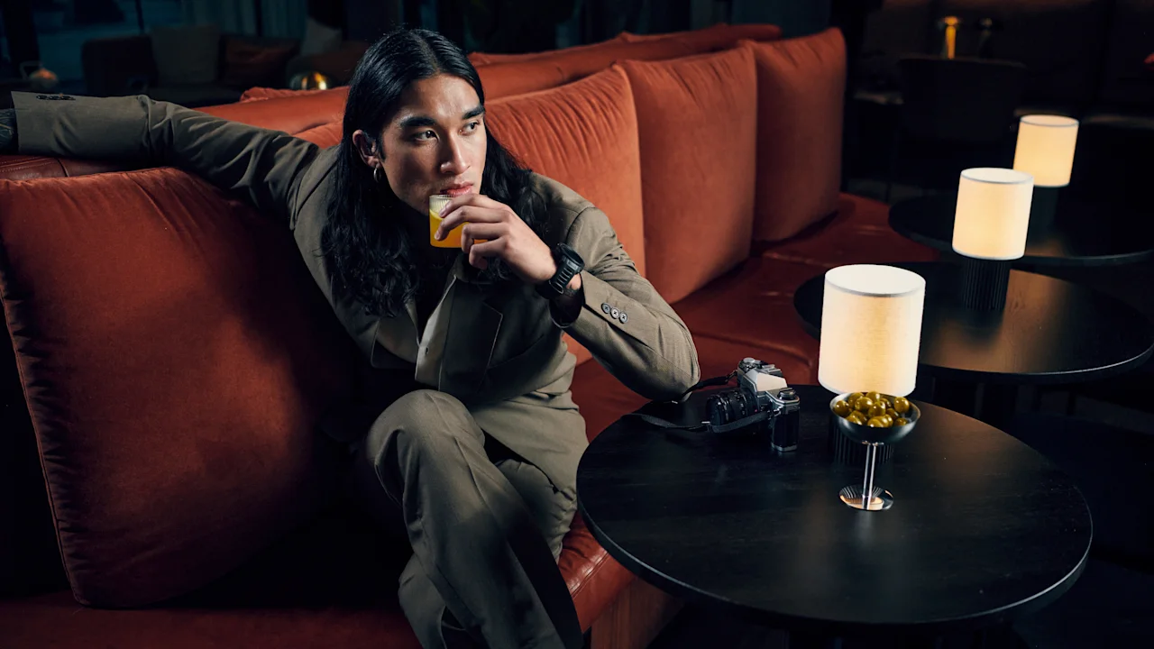 A person relaxing on a red couch in a dimly lit lounge, holding a drink and looking thoughtfully into the distance.