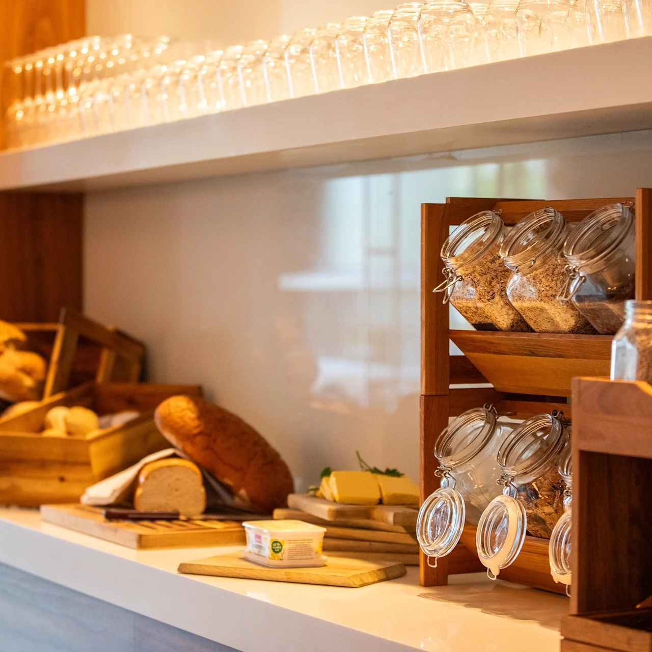 Kurver med assortert brød og stablede krukker med korn på en trehylle i en koselig kjøkkeninnstilling. Glass er stilt opp på øverste hylle.