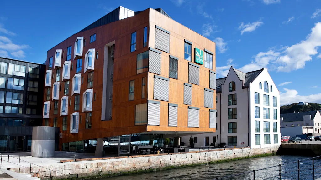 En moderne bygning i tre med utstikkende vinduer står ved en vannkant under en klar himmel. Ved siden av er en hvit bygning. Tekst: "Quality Hotel."