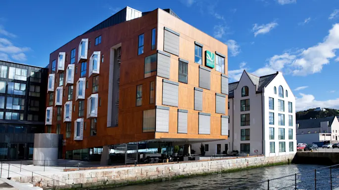 A modern wooden building with protruding windows stands by a waterfront under a clear sky. Adjacent is a white building. Text: "Quality Hotel."