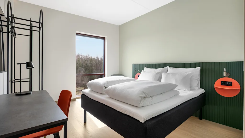 Et moderne hotellrom med en komfortabel seng, et skrivebord og et stort vindu som tilbyr en rolig utsikt over frodige trær.