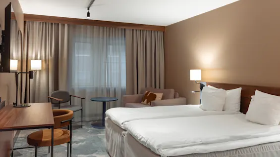 Et pænt arrangeret hotelværelse har to senge placeret side om side med hvidt sengetøj. I nærheden står et skrivebord med en stol over for et stort vindue, mens en sofa og lamper skaber en hyggelig atmosfære.