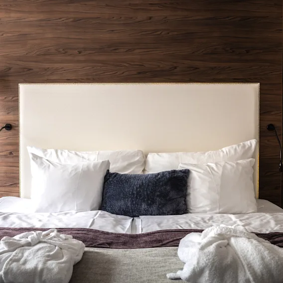 Ett mysigt hotellrum med en bekväm säng fylld med flera kuddar och utlagda morgonrockar, upplyst av stilrena överhängande lampor. De träfärgade detaljerna ger värme och elegans till rummet.