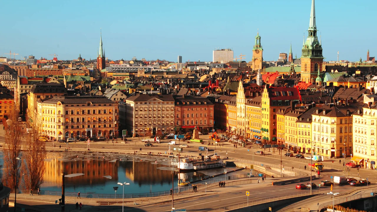 Fargerike historiske bygninger omgir et travelt bytorg med mennesker som går nær en reflekterende vannvei. Spir og tårn reiser seg mot en klar blå himmel og danner en pittoresk byscene.