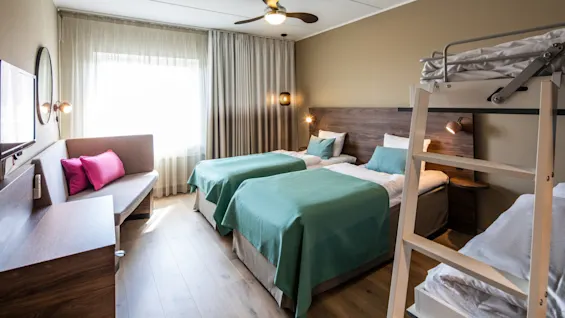 Three beds with green bedspreads are arranged in a neatly furnished bedroom. A ceiling fan spins above, and a sofa with pink cushions sits by a curtained window.