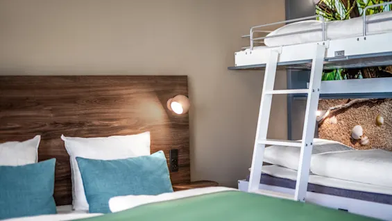 Bunk bed with white bedding stands next to a wooden headboard bed with green pillows in a softly lit room featuring beige walls and an image of an outdoor scene.