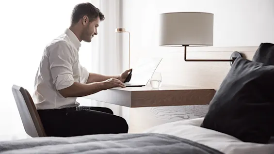 A man types on a laptop at a wooden desk in a modern room with a bed, lamp, and glass of water.