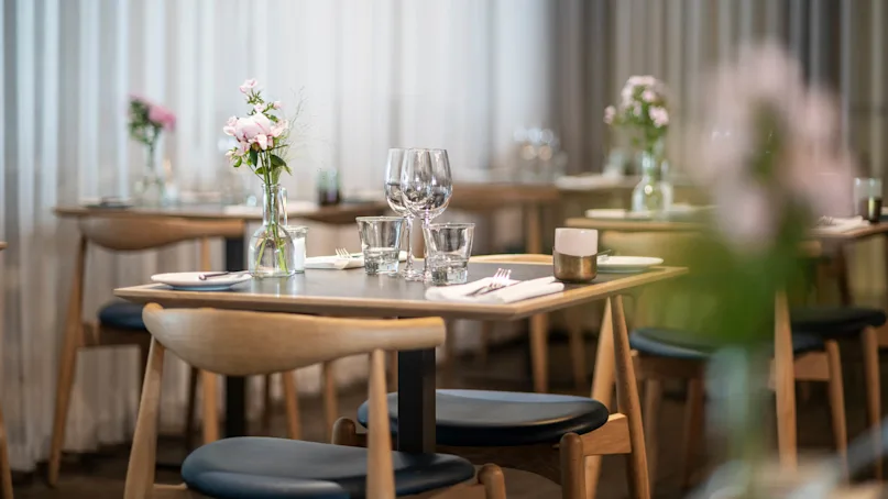 Table set with wine glasses, flowers in vase, and cutlery arranged neatly in a softly lit restaurant, surrounded by wooden chairs and sheer curtains.