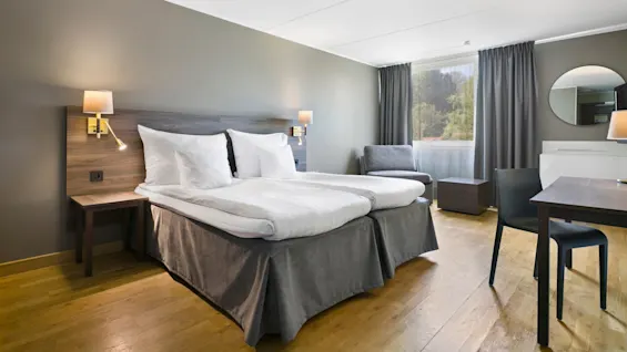 En seng med hvidt sengetøj står ved siden af et lille bord med lampe i et moderne hotelværelse. Stort vindue med gardiner og skovudsigt, stol og skrivebord i baggrunden.