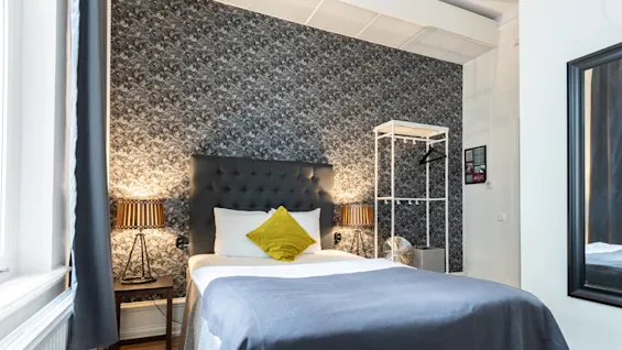 A neatly made bed with a dark headrest and a decorative pillow in a bedroom with elegant decor and luminous lamps.