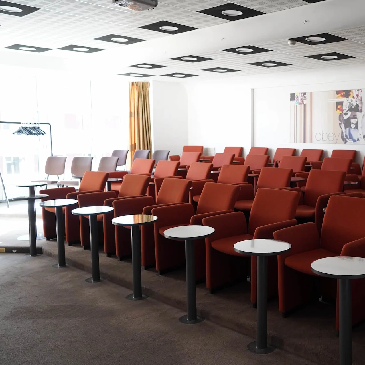 Red armchairs with circular tables are arranged in rows in a conference room. A wall features art with the words "obey dreams". Ceiling tiles are patterned.