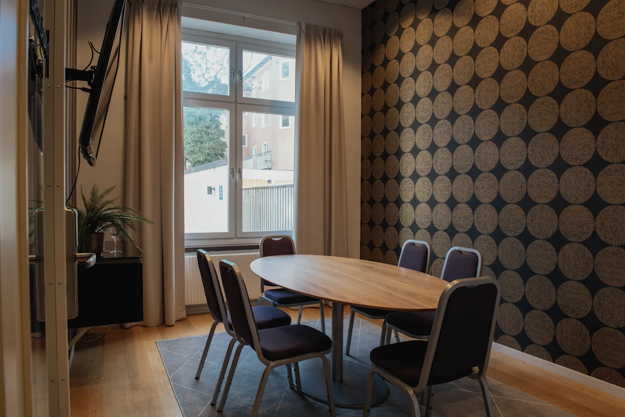 Ett modernt mötesrum med ett ovalt träbord, sex stolar, ett stort fönster och en väggmonterad TV. Tapeten har ett cirkulärt mönster.