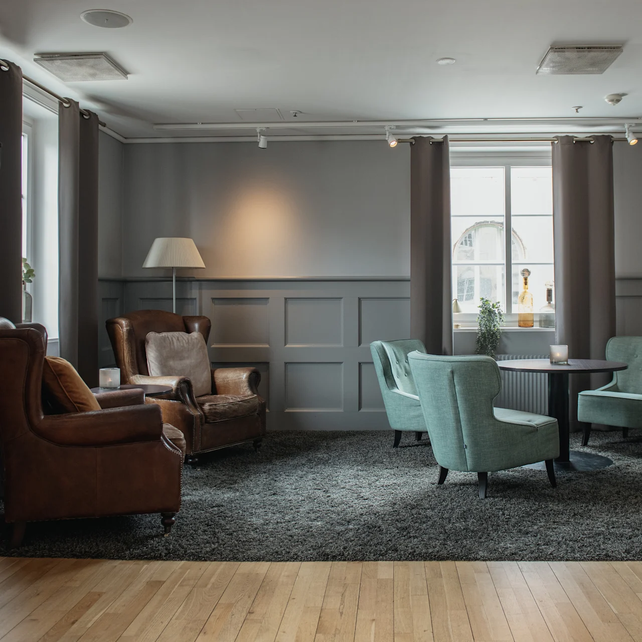 Brown leather armchairs and teal chairs surround a dark table on a grey rug. A guitar hangs on a wall, and a lamp stands nearby in an elegant room with wooden flooring.