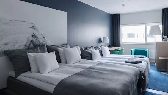 Ett modernt hotellrum med två prydligt bäddade sängar i grått och vitt, en stor landskapskonstverk ovanför, två turkosa stolar vid ett fönster och ett litet skrivbord med lampa.