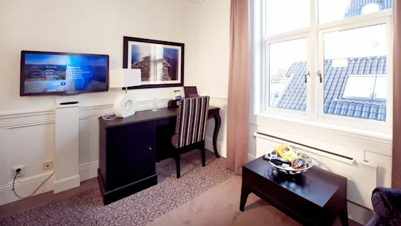 Ett modernt kontorsrum med en väggmonterad platt-TV, ett skrivbord med stol och fönster som vetter mot hustak.