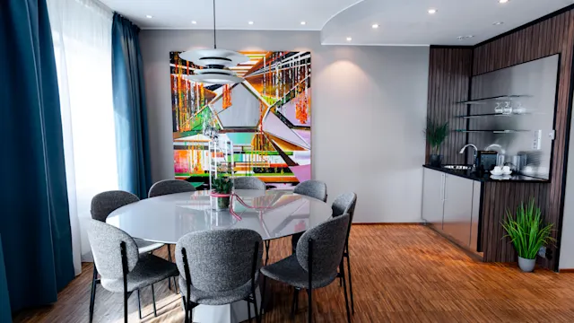 Dining table surrounded by gray chairs, set beneath a modern pendant light. In the background, a vibrant abstract painting hangs on a light gray wall, near a kitchenette and a window with curtains.