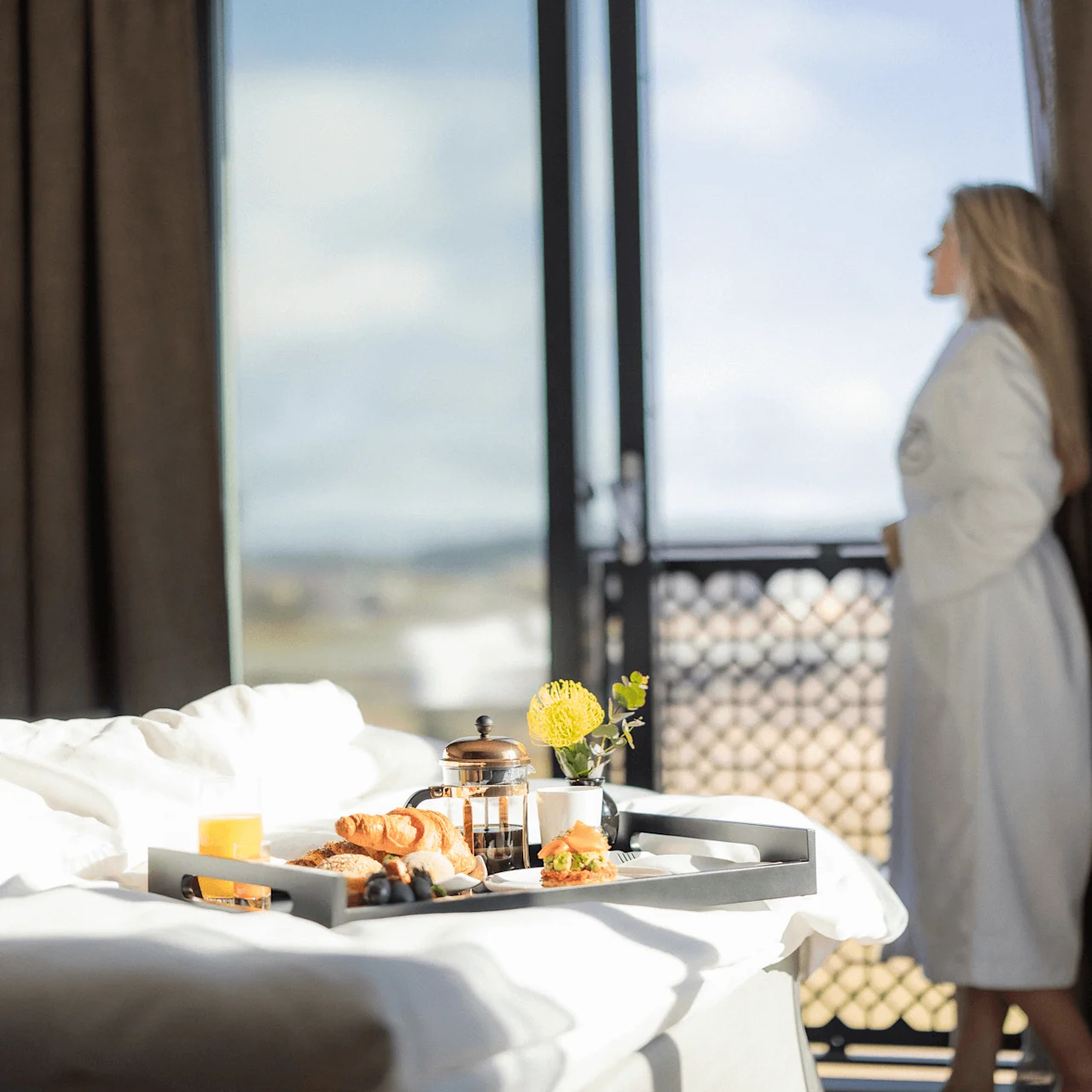 En frukostbricka på en säng med en suddig person i badrock som tittar ut genom ett soligt hotellrumsfönster.