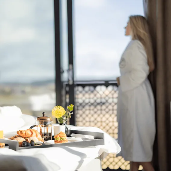En frokostbrett på en seng med en uskarp person i badekåpe som ser ut av et solrikt hotellromvindu.