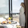 A breakfast tray on a bed with a blurred person in a bathrobe looking out of a sunny hotel room window.