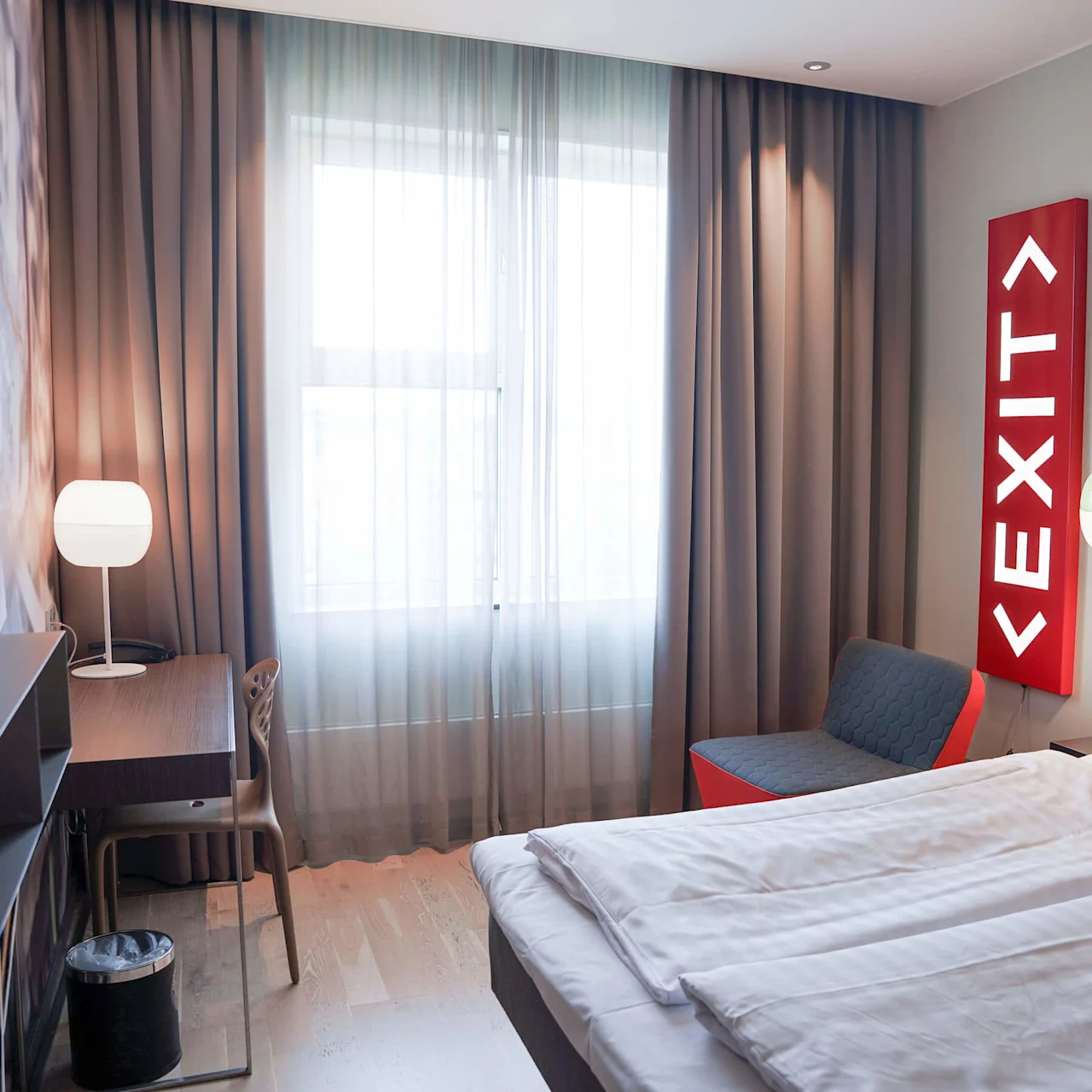Et minimalistisk hotellrom med en seng, stol, TV og en lampe. Et opplyst "EXIT"-skilt tilfører en quirky detalj mot en grå vegg.