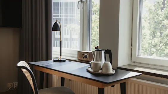 Et skrivebord har en lampe og et brett med en vannkoker og kopper. Sollys filtrerer gjennom store vinduer, som lyser opp det moderne rommet. Ingen tekst er synlig.