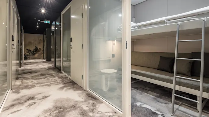 Kompakte sovebokser med køyesenger i en moderne setting. Korridoren har mønstrede tepper, opplyst av lys over, med tåkete skogbilde i enden.