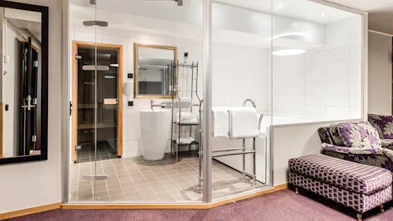 Ett modernt badrum med glasväggar intill en sittplats med en lila mönstrad soffa och matchande fotpall, speglar en lyxig inredningsdesign.