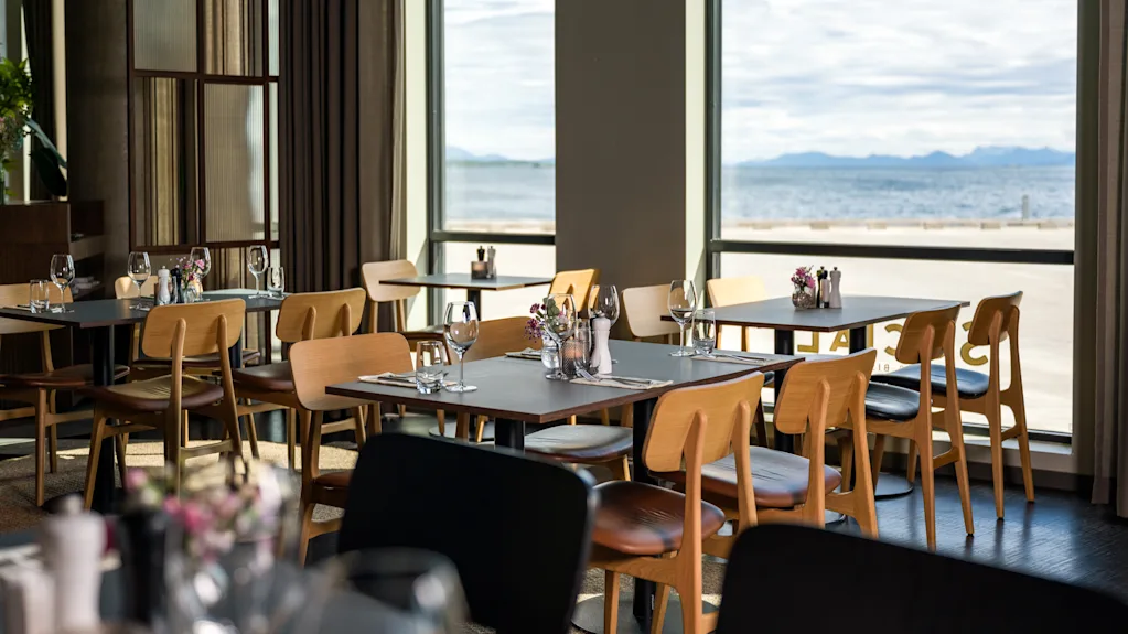 En lys restaurant på Quality Hotel Harstad med bord dekket for bespisning, med utsikt over vann og fjell.
