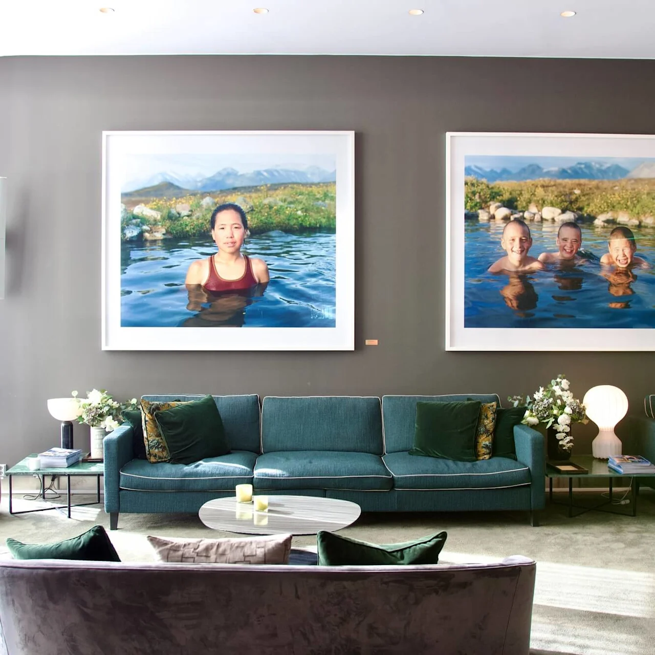 A modern lounge features teal sofas, a round table, and wall art of swimmers in serene water with mountainous backgrounds. Soft lighting and a floral arrangement enhance the cozy ambiance.