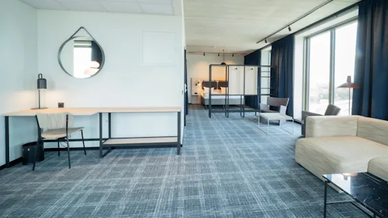 Tilava hotellihuone, jossa on moderni sänky, kirjoituspöytä tuoleineen ja oleskelualue sohvalla. Suuret ikkunat sinisillä verhoilla tuovat luonnonvaloa ja luovat ilmavan tunnelman.