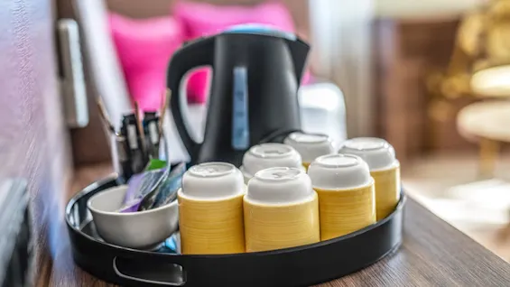 A tray holds six upside-down cups and a black electric kettle on a wooden surface, alongside stirrers and packets, in a cozy room with pink pillows in the background.