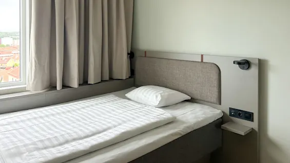 En pent redt enkelt seng med stripete sengetøy står ved siden av et stort vindu med beige gardiner i et minimalistisk rom med lyse vegger.