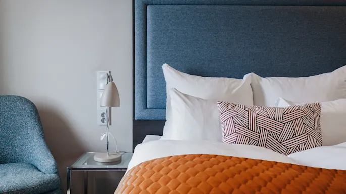 Bed with patterned pillows and orange quilt; adjacent lamp on nightstand; chair nearby. Room features neutral tones with a modern style.
