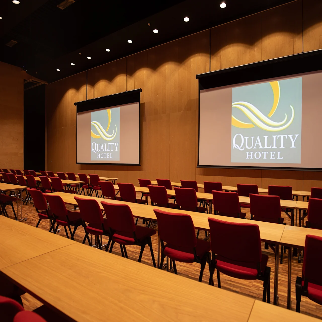 Skrivebord og røde stoler er arrangert i rader mot to store skjermer som viser "QUALITY HOTEL" i et moderne konferanserom med trevegger og varm belysning.