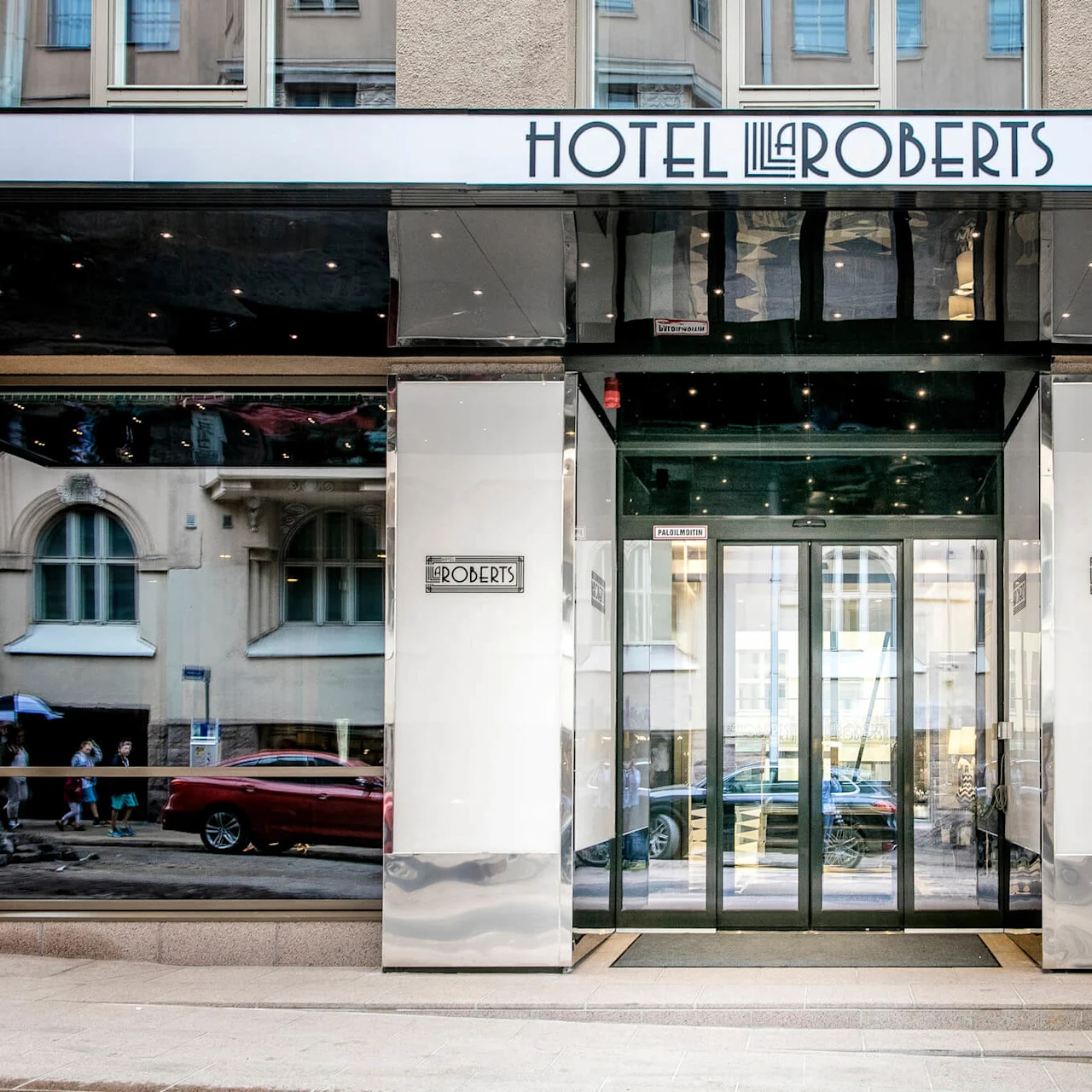 Inngang til Hotel Lilla Roberts med glassdører, som reflekterer den omkringliggende gaten. Skilt leser "Hotel Lilla Roberts", "ROOBERTS", og "PÄÄOVI MAIN ENTRANCE". Potteplanter rammer inn inngangen.
