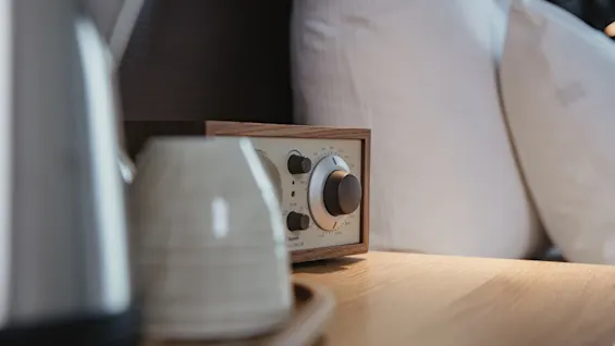 A small wooden radio sits on a wooden table, surrounded by pillows and a ceramic mug, creating a cozy bedroom atmosphere.