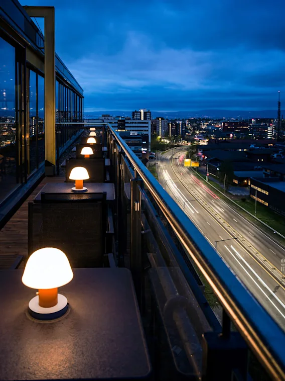 Utendørs terrassebord med glødende lamper står langs en høy balkong med utsikt over bylandskapet om natten. I det fjerne er ordet "TILIT" opplyst på en bygning.