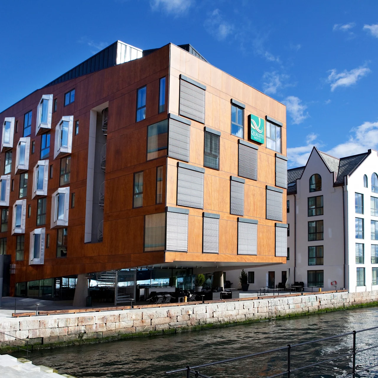 En modern träbyggnad med utskjutande fönster står vid vattnet under en klar himmel. Intill står en vit byggnad. Text: "Quality Hotel."
