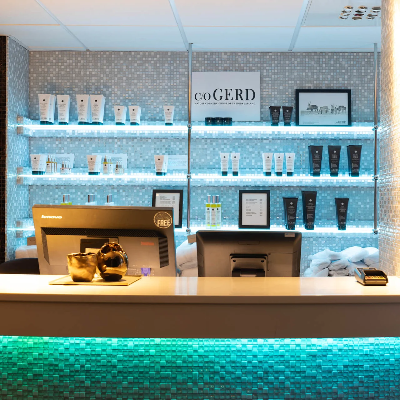 Datorbildskärmar står på en receptionsdisk, visar kosmetika på hyllor med mjuk belysning i en modern spamiljö. Text inkluderar: "C/O GERD NATURE COSMETIC GROUP OF SWEDISH LAPLAND".